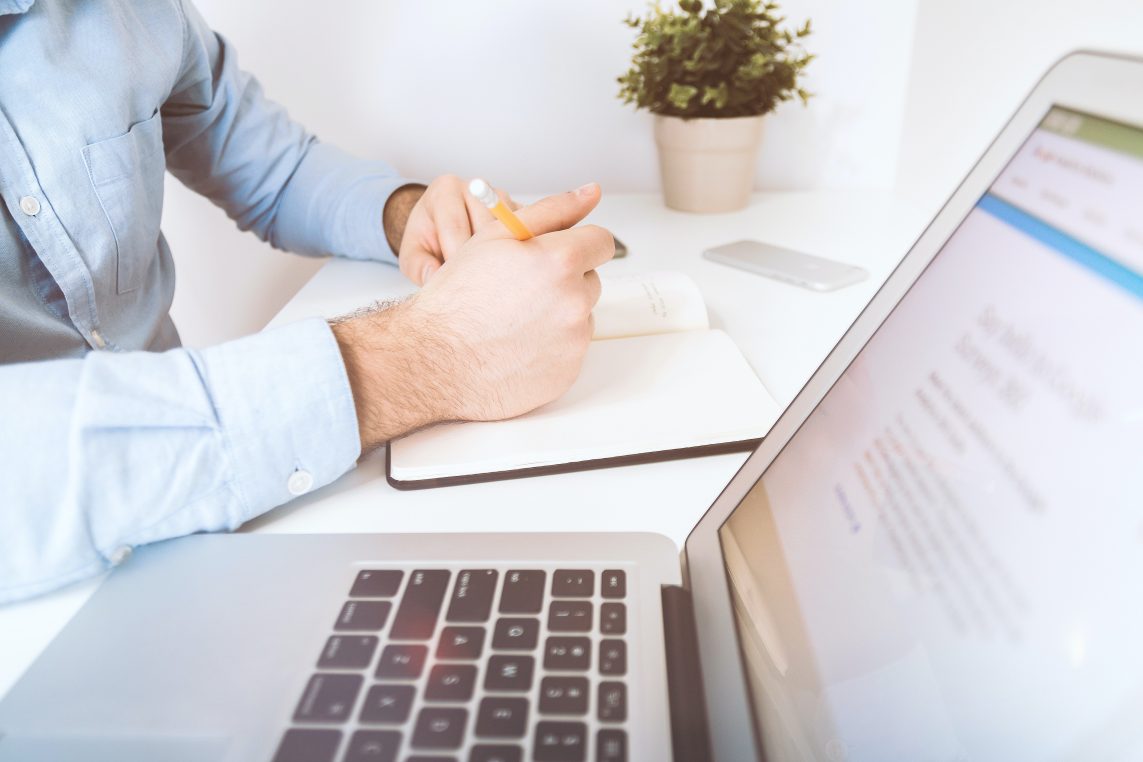 Person's hands write with a pencil in an open notebook while a laptop shows a blurred webpage (text unreadable) and a small potted plant sits on a white desk.