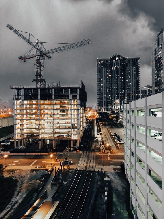 A construction crane looms over a lit, partially built multi-story structure; train tracks lead toward a distant high-rise along a wet city street at night under stormy clouds.