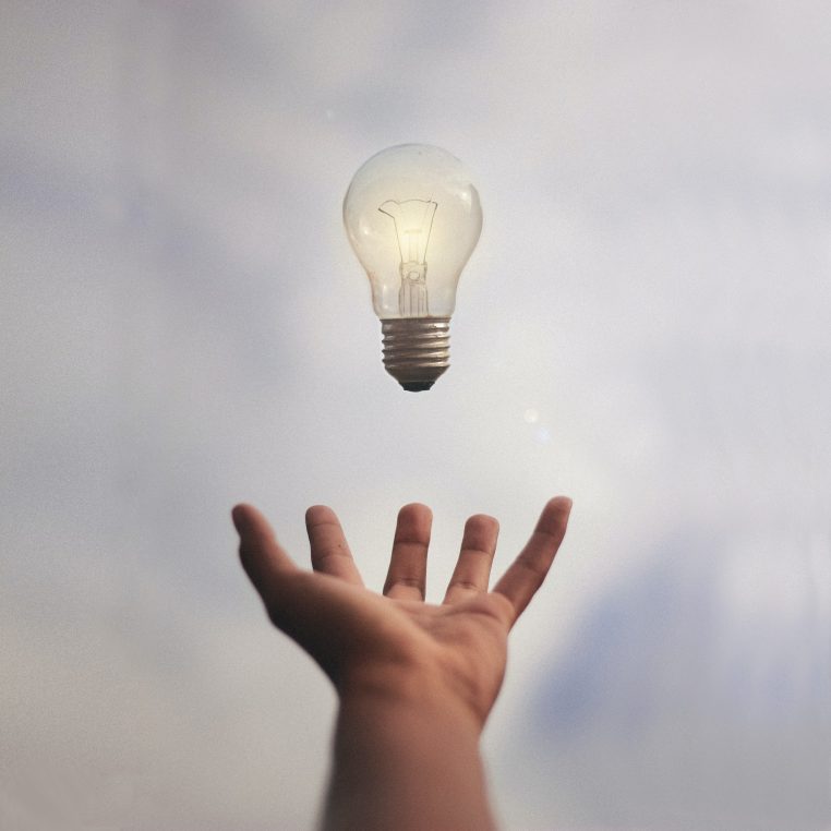 A clear incandescent light bulb floats above an outstretched hand against a soft, cloudy sky, suggesting levitation or offering; the filament is faintly glowing.