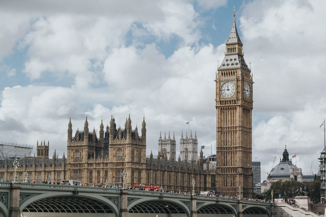 Elizabeth Tower (Big Ben) rises beside the Gothic Houses of Parliament; clock faces visible, overlooking Westminster Bridge crowded with pedestrians and red buses under a cloudy sky.