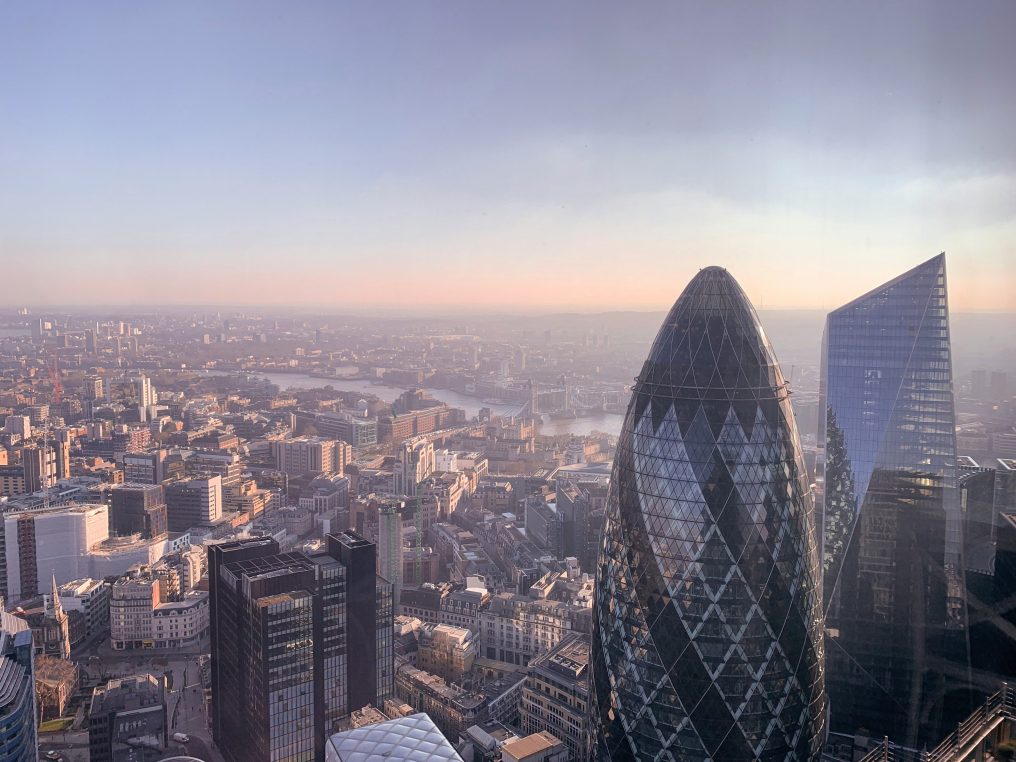 Egg-shaped glass skyscraper rises above surrounding high-rises as soft sunrise light bathes a sprawling cityscape with a river and distant low-rise neighborhoods under a pale sky.