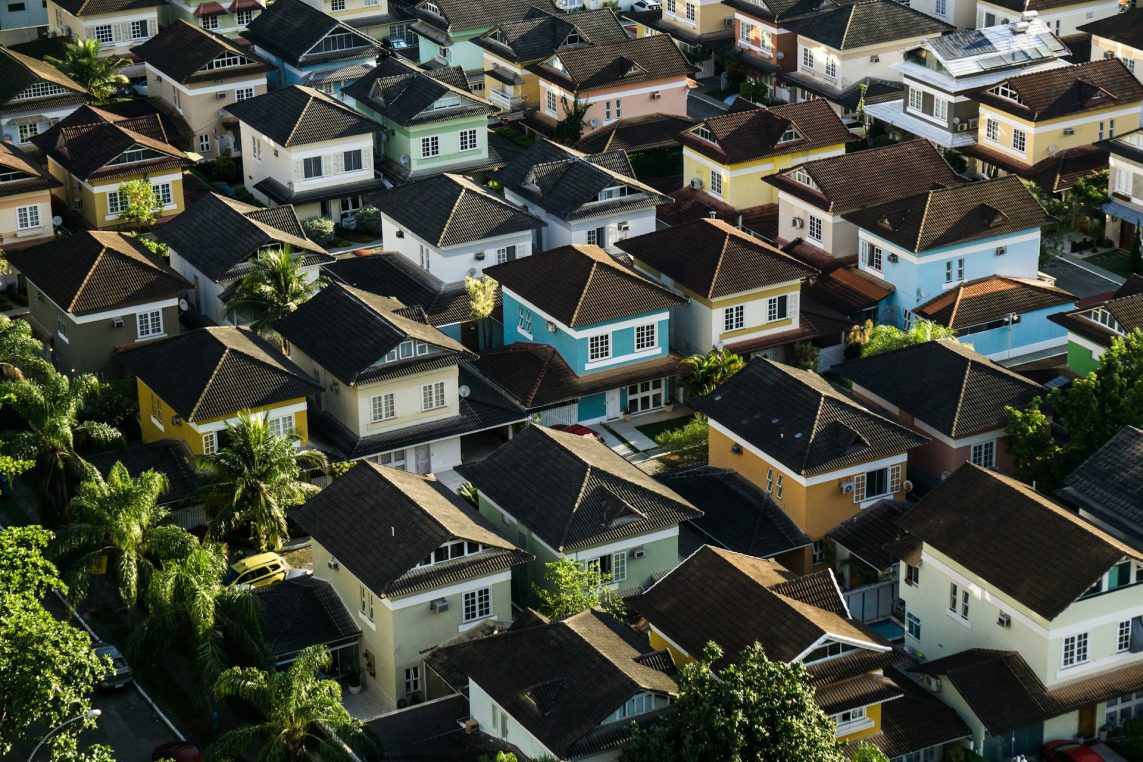 Rows of colorful two-story houses with dark tiled roofs sit closely packed, forming a dense suburban neighborhood punctuated by palm trees and narrow driveways in late-afternoon sunlight.