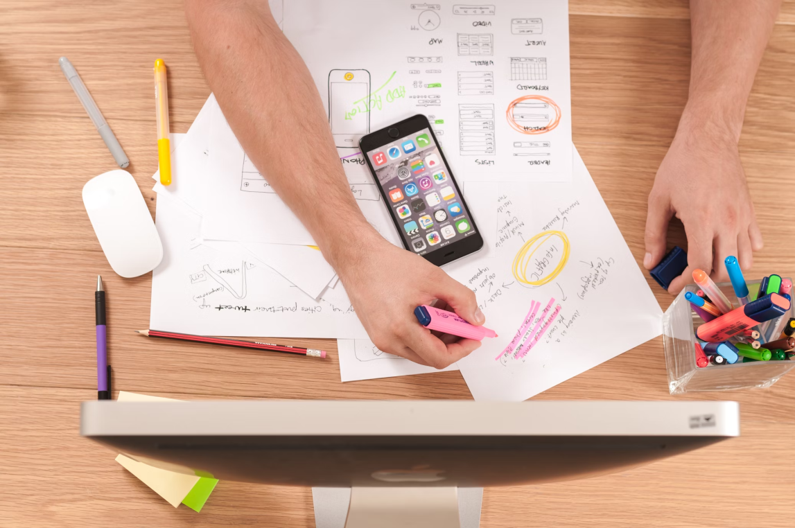 Person's hands highlighting notes with a pink highlighter over sketched wireframes; a smartphone rests on papers amid pens, markers, a mouse, and an iMac on a wooden desk.