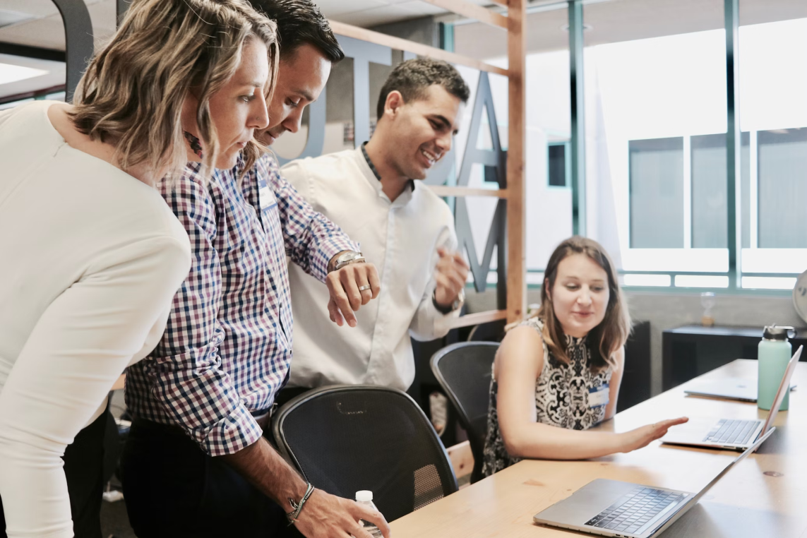 Four colleagues lean toward a laptop; a seated woman gestures at the screen while three teammates stand watching in a bright open-plan office with windows, shelving, and a wooden table.
