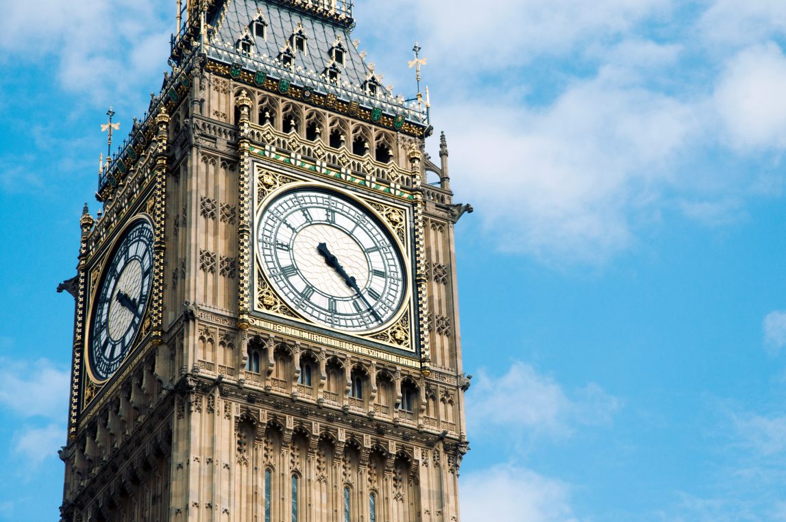 An ornate clock face on Elizabeth Tower displays time, its black hands pointing roughly to eleven and two; gilded Gothic stonework rises against a clear blue sky.