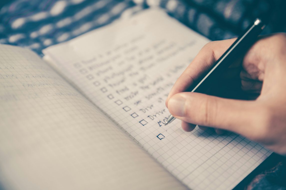 Right hand holds a pen checking items on a handwritten checklist of square boxes in a grid notebook, pages spread on a soft patterned surface; handwriting is blurred and illegible.
