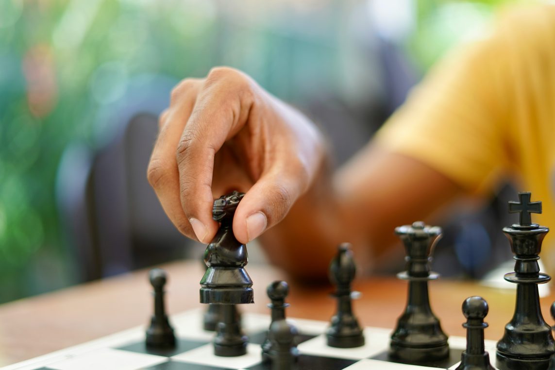 A hand lifts a black knight chess piece over a checkered board; other black pieces stand nearby, with a blurred green background and a person wearing a yellow shirt.