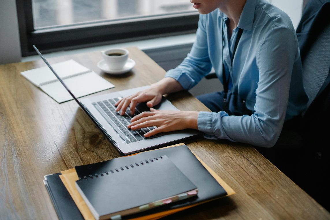 Person's hands typing on a laptop keyboard at a wooden desk, with stacked notebooks and folders, a coffee cup nearby, and a windowed office background.