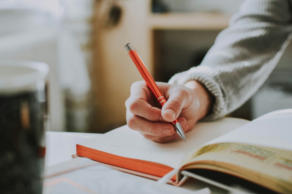 Hand holding a red pen writes in a notebook; an open book lies nearby and a blurred mug sits on the desk, sweater-clad arm resting in a warm indoor workspace.