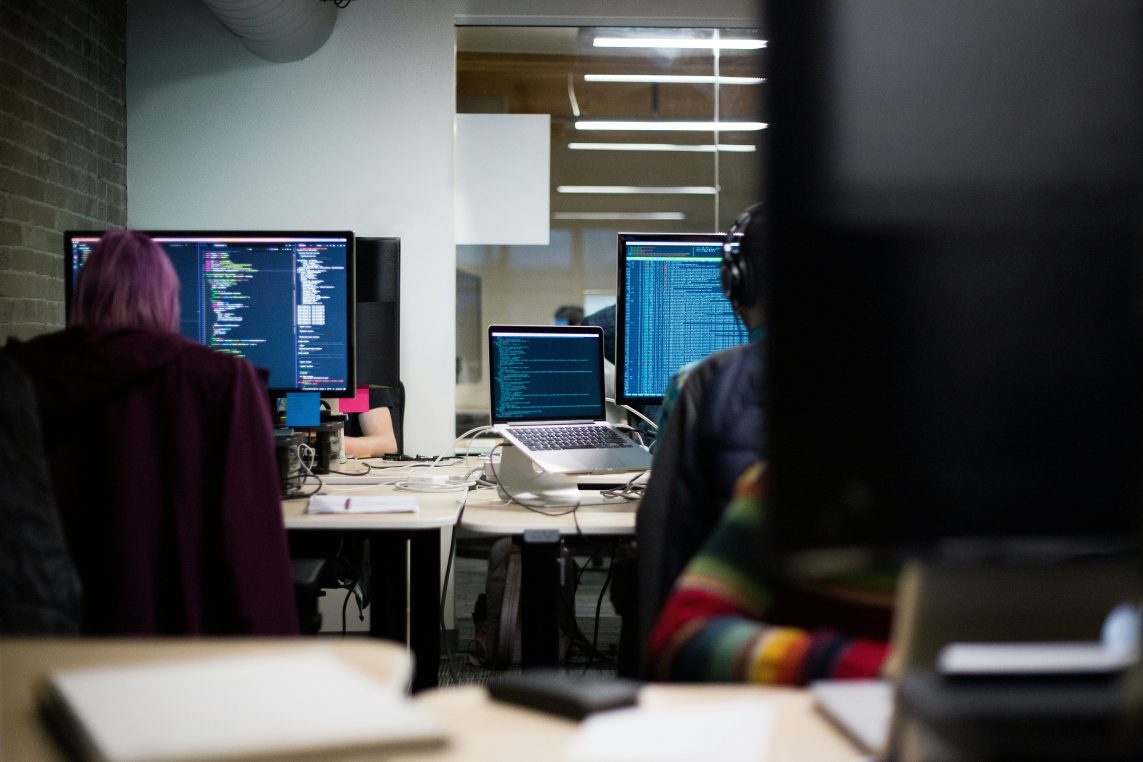 Multiple computer monitors and a laptop display lines of code while programmers work at desks; cables, headphones and a glass meeting room in a dim open-plan office. On-screen text unreadable.