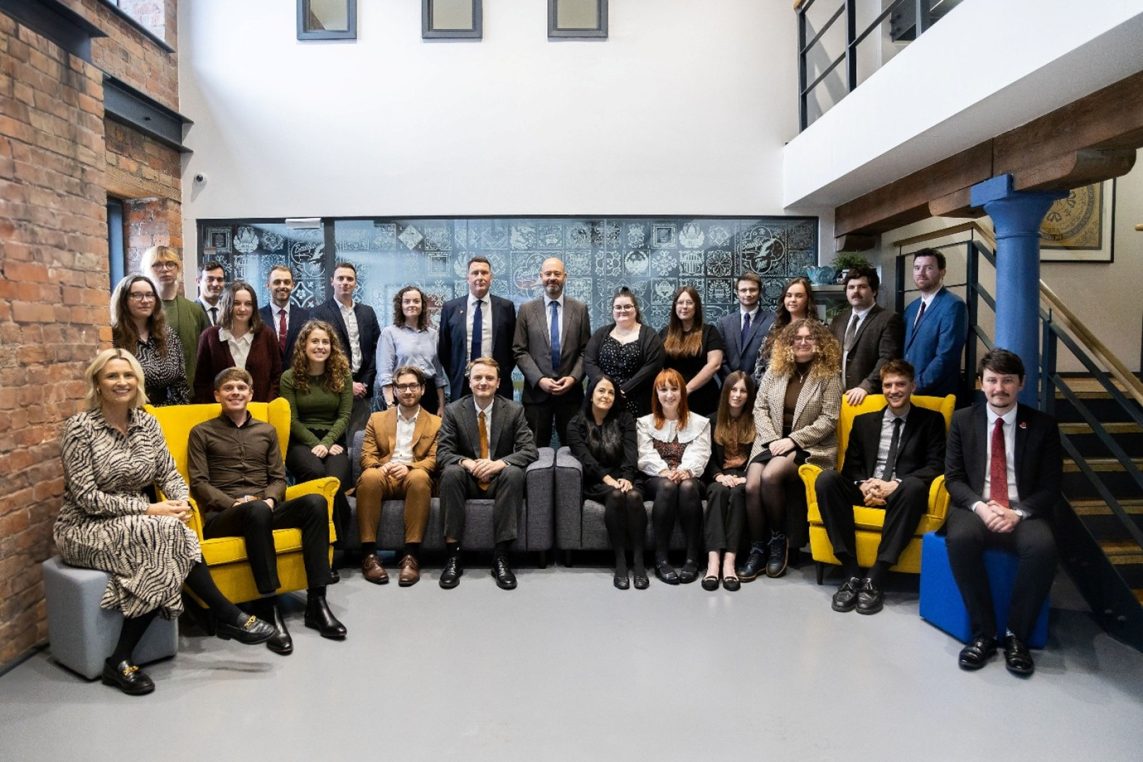 A group of about twenty professionally dressed adults sit and stand in two rows, posing for a formal group photo inside a modern industrial atrium with exposed brick, a mural, and a staircase.