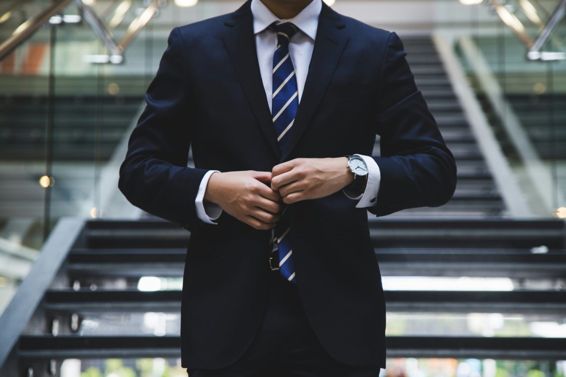 A man in a navy blue business suit, with his hands held next to the jacket buttons as if he is about to open it