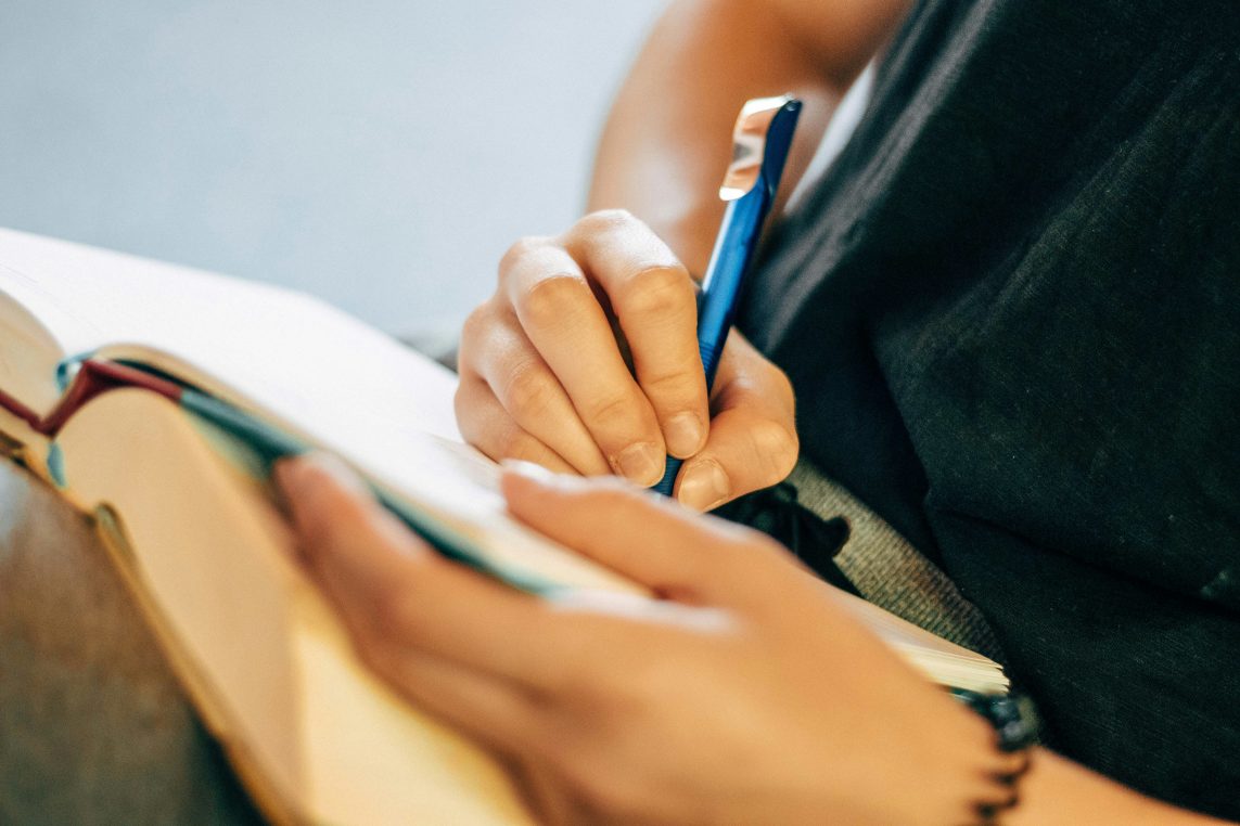Pair of hands holding a blue pen, writing on an open notebook; other hand steadies the pages on a lap, casual indoor setting with dark clothing and soft blurred background.