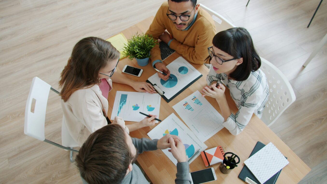 Four coworkers gather around a wooden table, pointing at printed pie charts and graphs, discussing data; smartphones, notebooks and a small potted plant sit on the table.