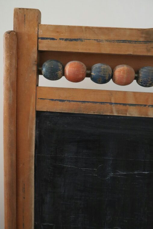 Wood-framed chalkboard panel with a row of worn painted wooden beads sliding on a top rod; scuffed black writing surface and distressed honey-colored frame suggest a vintage classroom.