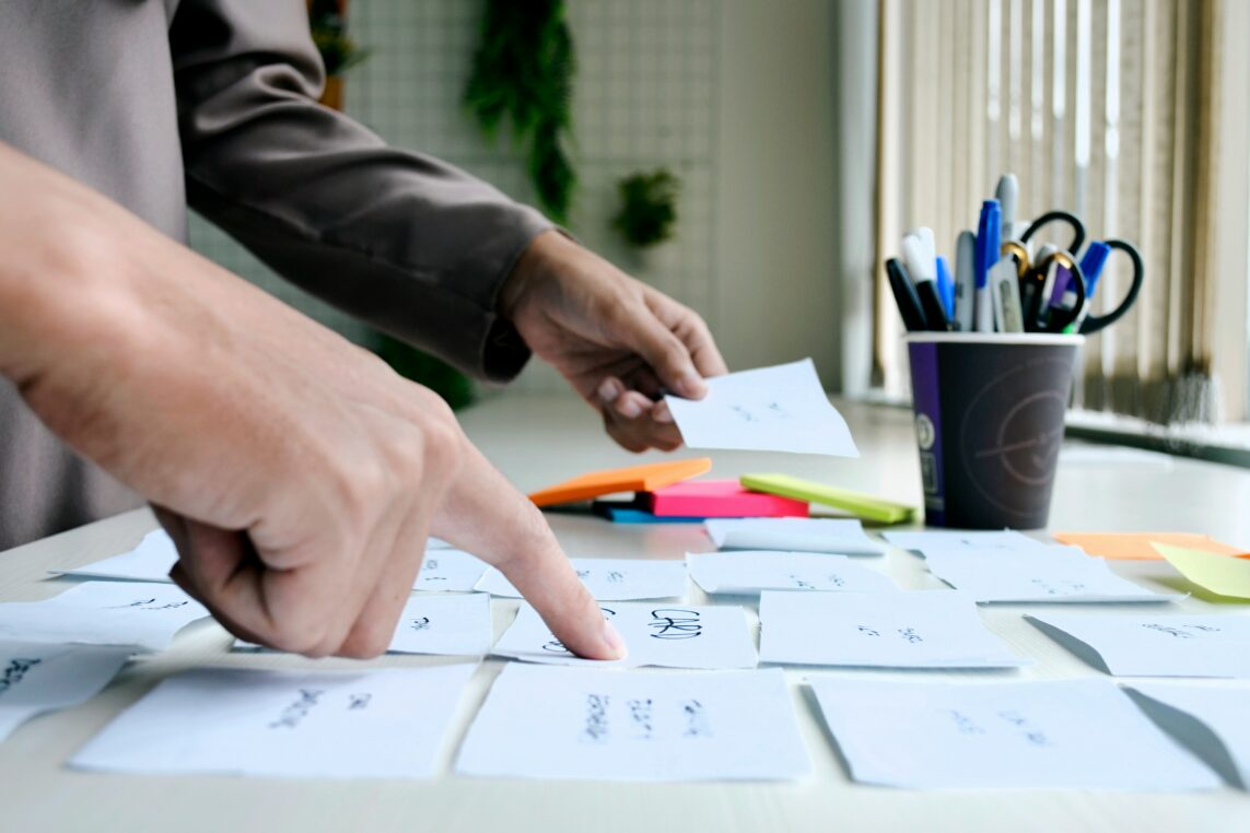 Two hands sort and point at scattered sticky-note cards on a white desk, one holding a card; colorful highlighters and a pen-filled cup sit by window blinds in an office-like space.